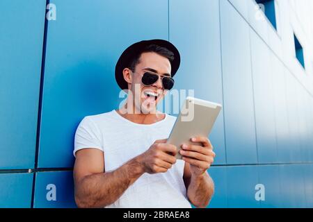 Schöne fröhliche Mann an Tablet Bildschirm schaut, fröhlich lachend, etwas zu beobachten. Im weißen T-Shirt, gekleidet in schwarzen Hut und Sonnenbrille. Im Freien Stockfoto