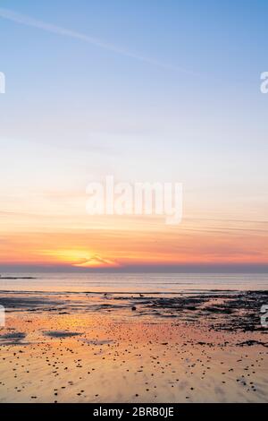 Der Morgenhimmel über der Nordsee vom Viking Bay Beach in Broadstairs. Ebbe, niedrige Sicht mit sanften Wellen, die an der Küste plätschern, sehr ruhiges Meer mit einer Schicht von trüben orangen Wolken am Horizont und darüber ein überwiegend klarer blauer Himmel. Stockfoto