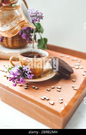 Goldene Porzellan Teekanne gießt einen Strom Tee in Tasse auf Holztablett mit Herzen mit Schokoladenkuchen, lila lila Blüten in einem Weidenbasen dekoriert dekoriert Stockfoto