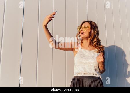 Schöne lockige Mädchen in der Sonnenbrille ein selfie auf Telefon, stehend gegen die weiße Wand, im Freien. In modisch gekleidet. Stockfoto