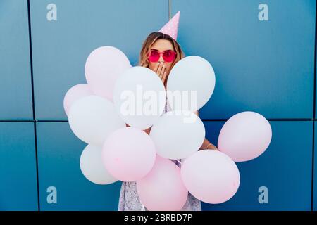 Schöne junge Mädchen schaut überrascht beim Stehen mit Luftballons. Tragen rosa Sonnenbrille und Hut, außerhalb. Stockfoto