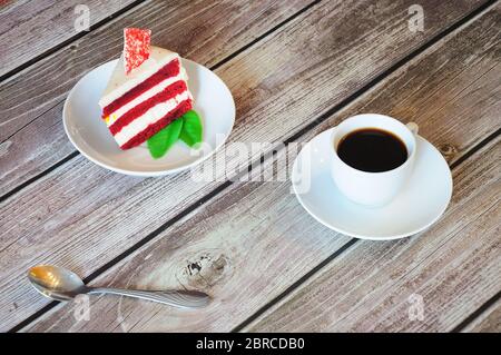 Ein Stück roter Samtkäsekuchen mit einem Minzblatt und einer Tasse frischen Cappuccino steht auf einem Holztisch. Nahaufnahme. Stockfoto