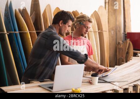 Zwei kaukasische männliche Surfboard-Hersteller arbeiten an Projekten und mit einem Laptop-Computer mit Surfbrettern Stockfoto