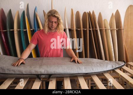 Kaukasischer männlicher Surfboard-Hersteller, der ein Surfbrett mit Surfbrettern in einem Rack im Hintergrund sieht Stockfoto