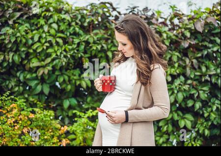 Eine schwangere junge Frau, die an der Hecke steht und aus einer Tasse trinkt. Schwangere Frau, die sich im Park entspannt. Stockfoto