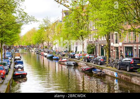 Amsterdam, Niederlande - 27. April 2019: Kleines Boot auf dem Kanal im Stadtzentrum der niederländischen Hauptstadt. Parkende Autos an grünen Bäumen entlang des Kanals. Horizontales Foto. Stockfoto