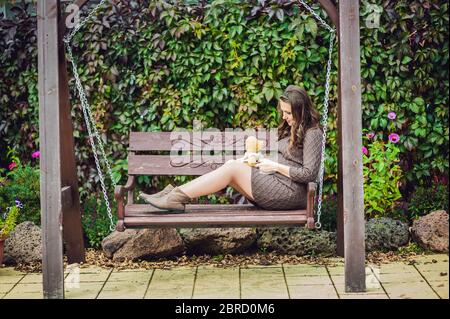 Eine schwangere junge Frau auf einer Schaukel, mit einem Teddybär. Schwangere Frau im Park entspannen. Stockfoto