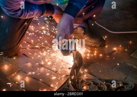 Vorderansicht der Hände eines Mannes, der an einem Metallteil einer Gartenbank arbeitet, mit einem elektrischen Schleifer, während Funken in der industriellen Arbeit herumfliegen Stockfoto