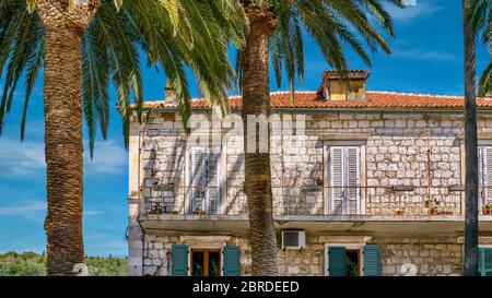 Die Fassade einer alternden Villa in Kroatien an einem Sommernachmittag, mit hölzernen Türen, Balkon, Steinmauern, Terrakotta-Fliesen und Palmen. Stockfoto