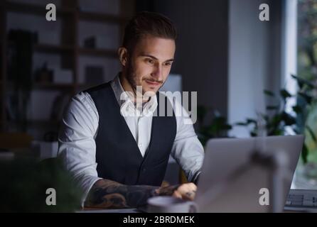 Ein Geschäftsmann mit Laptop sitzt im Büro drinnen am Schreibtisch und arbeitet spät. Stockfoto