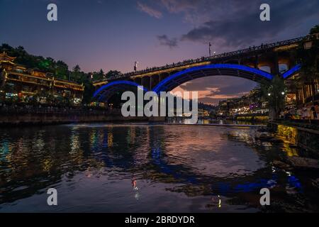 Feng Huang, China - August 2019: Nach Sonnenuntergang Nacht Blick auf die Straßenbrücke über den Tuo Jiang Fluss und Holzhäuser in der alten Altstadt von Fenghuang Stockfoto