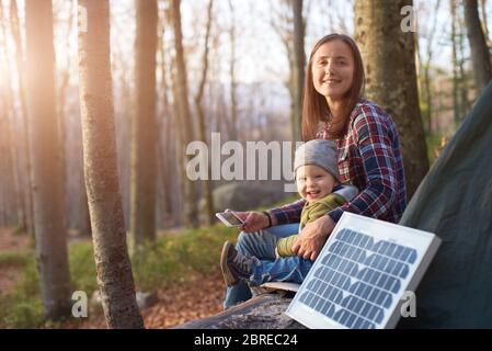 Tourist Solarpanel im Vordergrund einer jungen Familie im Wald. Das Baby und die Mutter sitzen in den Sonnenstrahlen und schauen mit einem Lächeln auf die Kamera. Frau mit einem Mobiltelefon Stockfoto