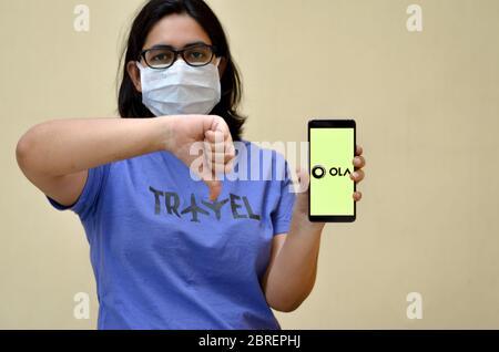 New Delhi, Indien, 2020.Mädchen trägt Maske zeigt Ola App auf ihrem Handy-Bildschirm mit Daumen nach unten vor gelbem Hintergrund.Es ist ein Taxi Aggregator Stockfoto