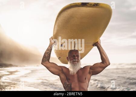 Happy Fit Senior mit Spaß beim Surfen bei Sonnenuntergang - sportliches bärtiger Mann Training mit Surfbrett am Strand Stockfoto