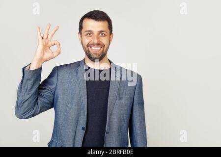 Gut aussehende, selbstbewusste junge Mann garantiert einwandfreie Qualität, zeigt eine gut bestätigende Geste, lächelt, zufrieden, genehmigt eine gute Idee, empfiehlt eine Promo stimmt mit Ihnen steht auf einem weißen Hintergrund Stockfoto