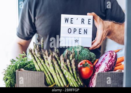 Ende der Sperrung der Quarantäne. Mann hält Leuchtkasten mit Grußtext-Nachricht Wir sind offen und frische Gemüse und Gemüse-Box. Einladender Lebensmittelgeschäft cli Stockfoto