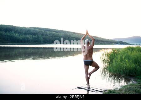 Rückansicht des älteren Mannes in Badeanzug, der im Freien am See beim Yoga steht. Stockfoto