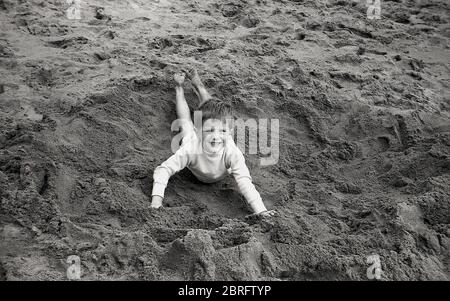 1961, historischer, glücklicher Junge, der in einem flachen, breiten Loch oder Loch liegt, das er gerade am Sandstrand gegraben hat, England, Großbritannien. Stockfoto