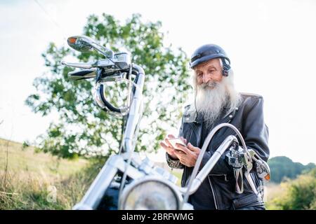 Senior-Mann mit Motorrad in der Landschaft, mit Smartphone. Stockfoto