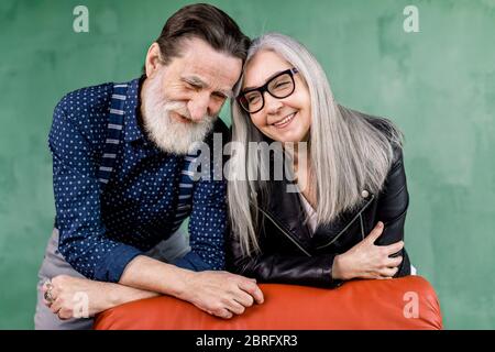 Lächelndes, wunderschönes Seniorenpaar, das die Zeit zusammen genießt, sich auf einen roten Stuhl stützt und die Stirn berührt, während man im modernen Zimmer vor sich steht Stockfoto