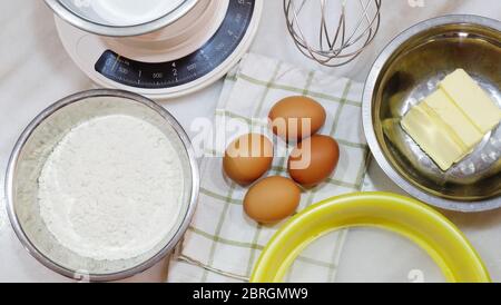 Backzutaten aus braunen Eiern, Weißmehl, Butter, Mehlsichter, Küchenwaage und einem Metallbesen, auf einer Tischplatte platziert. Stockfoto