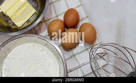 Backzutaten aus braunen Eiern, weißem Mehl, Butter und einem Metallbesen, auf einer Tischplatte platziert. Stockfoto
