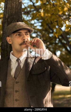 Retro-1920er-Porträt eines englischen Gangsters mit flacher Mütze. Auf der Straße raucht man eine Zigarette. Stockfoto