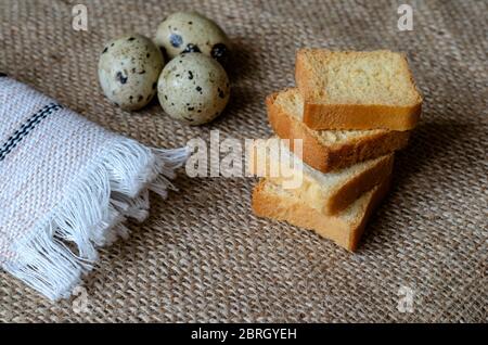 Quadratische Croutons und rohe Wachteleier auf Burlap. Vier kleine Croutons und drei Eier. Stillleben im ländlichen Stil. Ansicht von oben in einem Winkel. Selektiver Fokus Stockfoto