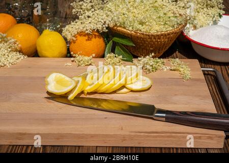 Auf einem Schneidebrett liegt ein Küchenmesser und eine in Scheiben geschnittene Bio-Zitrone, im Hintergrund sieht man Orangen, Zitronen und andere Zutaten Stockfoto