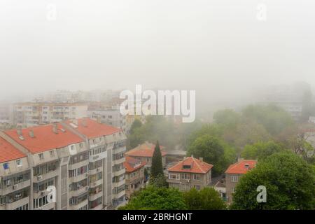 Wohnhäuser und Straßen mit grünen Bäumen in dichtem Nebel am Sommertag. Dichter Nebel kam aus dem Meer und bedeckte die Stadt. Stockfoto