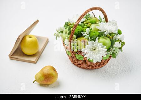 Stillleben mit einem Korbkorb gefüllt mit Früchten, weißen Blumen und einem offenen Buch. Stockfoto
