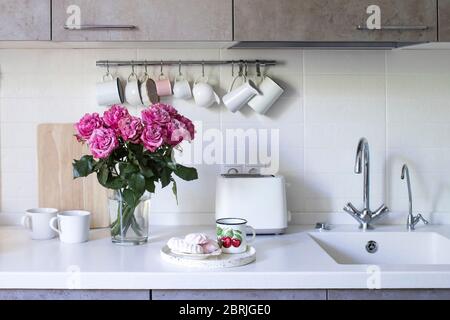 Küche in beigefarbenen Tönen. Becher und Milchkännchen hängen an Haken. Waschbecken mit Wasserhähnen. Bouquet von rosa Rose in einer Glasvase auf weißem Hintergrund auf einem Holztisch Stockfoto