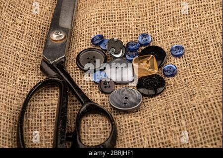 Mehrere mehrfarbige Knöpfe in verschiedenen Größen und große Vintage Sartorial Schere vor einem Hintergrund von grobtexturierten Burlap. Nahaufnahme Stockfoto