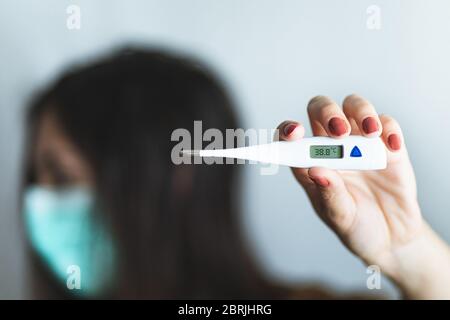 Vorderansicht der Hand einer Frau, die ein digitales Thermometer hält, das über 38 Grad Fieber anzeigt. Hintergrund der kranken Frau, die eine chirurgische Maske trägt, besorgt Stockfoto