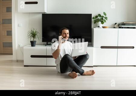 Mann mit Bart, zu Hause mit einem Laptop vor ihm arbeiten und mit einem Freund oder Geschäftspartner oder Kunden mit einem Smartphone und sprechen Stockfoto