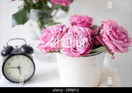 Ein Strauß rosa Rosen in einem Zinn weißen Eimer auf einem alten Hocker mit Wecker und einer Serviette mit Stickerei neben dem Bett auf einem beigen Hintergrund Stockfoto