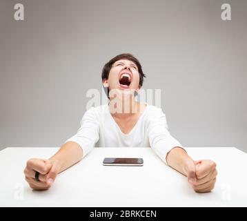 Frustrierte Frau, die einen temperamentvoller Wutanfall wirft, ihre Fäuste zusammenklammend und ein Handy vor sich auf dem Tisch anschreiend Stockfoto