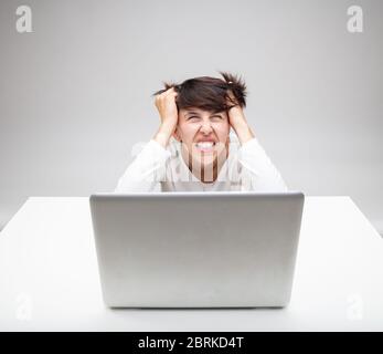 Frustrierte Geschäftsfrau zerreißt sich verärgert an den Haaren, als sie vor grauem Hintergrund an einem Laptop auf einem weißen Schreibtisch sitzt Stockfoto