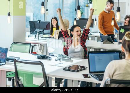 Fröhliche Angestellte jubelt, während sie ihre Hände hoch in die Luft hebt, während sie neben ihren Kollegen in einem modernen Büro sitzt. Stockfoto