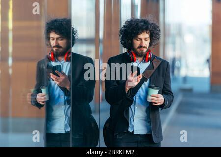 Fröhlicher Student mit Afro Haircut, der auf dem Campus spazieren geht, während er seine Mantasche und seine Kopfhörer trägt. Stockfoto