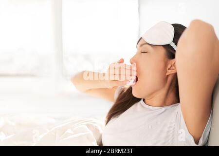 Früh morgens bei Sonnenaufgang aufwachen glücklich Frau gähnend sich im Bett ausstrecken. Schlafen zu Hause Frau Aufwachen in Morgen Sonnenlicht Stretching glücklich nach guter Nachtruhe Gefühl ausgeruht tragen Augenmaske. Stockfoto