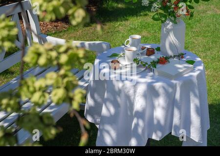Weiße Tischdecke gedeckter Tisch mit Kaffee, Kuchen, Buch und Vase im Garten durch eine weiße Bank hinter einem Baumzweig mit frischen grünen Blättern. Die Konz Stockfoto