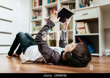 Teenager-Junge trägt Kopfhörer auf dem Boden liegen Spiel auf Tablet zu Hause spielen. Junge entspannen durch Online-Spiel auf digitalen Tablet. Stockfoto