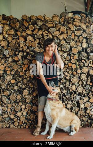 Lächelnde Frau mittleren Alters in Schürze mit Hund auf Haufen Der gesägten Protokolle Stockfoto