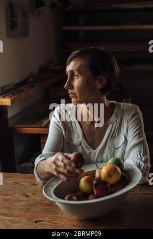 Frau mittleren Alters sitzt am Tisch mit Schale mit Früchten Stockfoto