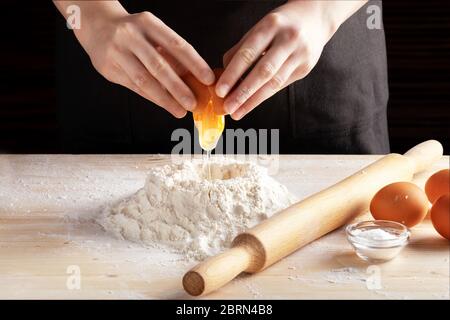 Vorderansicht der weiblichen Hände, die ein Ei in Mehl brechen, Teig mit einem hölzernen Nudelholz machen. Schritt-für-Schritt-Rezept für Knödel oder Knödel, Schritt 2. Stockfoto