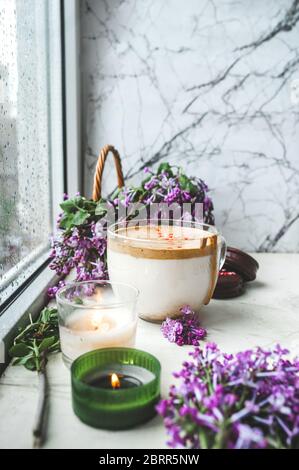 Dalgona Kaffee mit Schaum, Schokoladenkekse, brennende Kerzen und frische, saftige lila-lila Blüten am nassen Fenster mit Tropfen und Tropfen vom Regen. Stockfoto