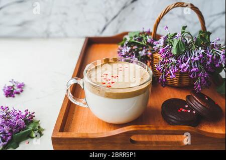 Holztablett mit Dalgona Kaffee mit Schaum, Schokolade Cookies, frische üppige lila lila lila Blumen in einem Weidenkorb. Gemütlich schönes Frühstück Stockfoto