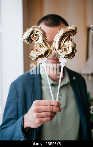 Der Mann in einer blauen Jacke hält kleine verfoldete Goldballons der Zahl 36 auf Stöcken in den Händen und bedeckt damit sein Gesicht. Sechsunddreißig Geburtstag. T Stockfoto