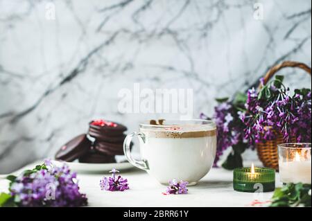 Dalgona Kaffee mit Schaum, Schokolade Cookies, brennende Kerzen und frische, üppige lila-lila Blumen auf einem weißen Marmorgrund. Gemütliche schöne Breakfas Stockfoto